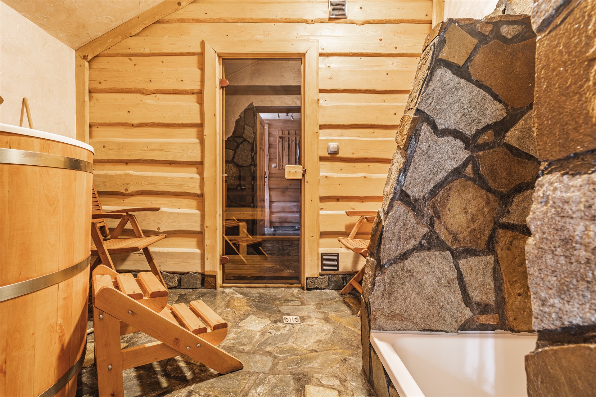 Przytulne wnętrze sauny z drewnianą balią, składanymi ławkami, kamiennym kominkiem i szklanymi drzwiami do sauny