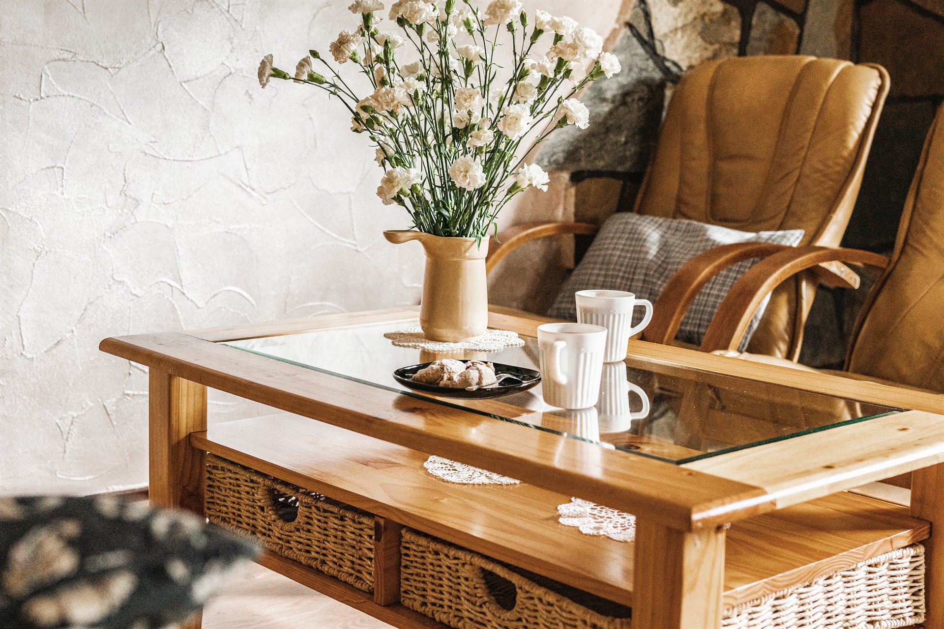 Drewniany stolik z szklanym blatem, wazon białych goździków, dwa białe kubki i talerz z ciastkami