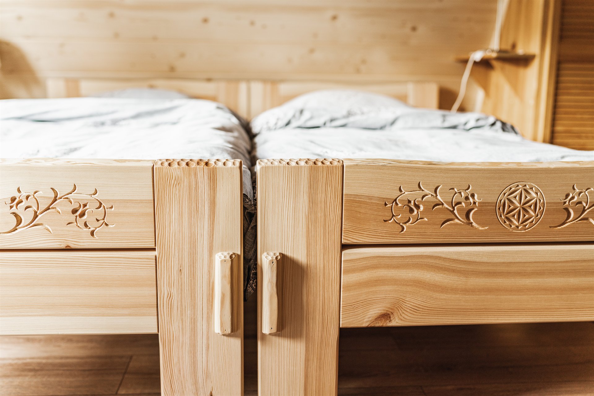 Dwa drewniane łóżka z jasnego drewna z rzeźbionymi motywami roślinnymi i okrągłym ornamentem, jasne pościele