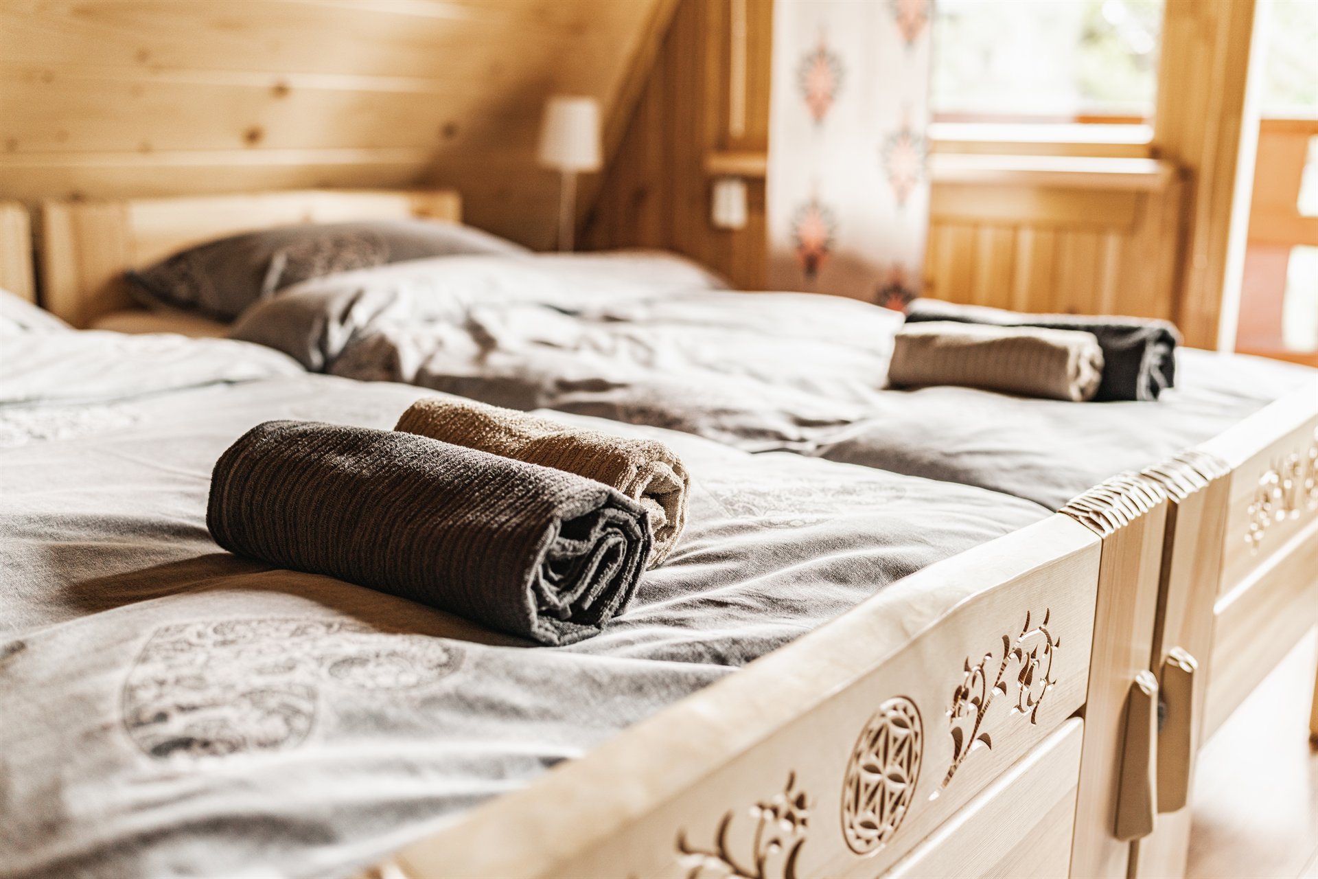 Podwójne łóżko w drewnianym pokoju z ozdobnym wezgłowiem i zrolowanymi ręcznikami na szarej pościeli