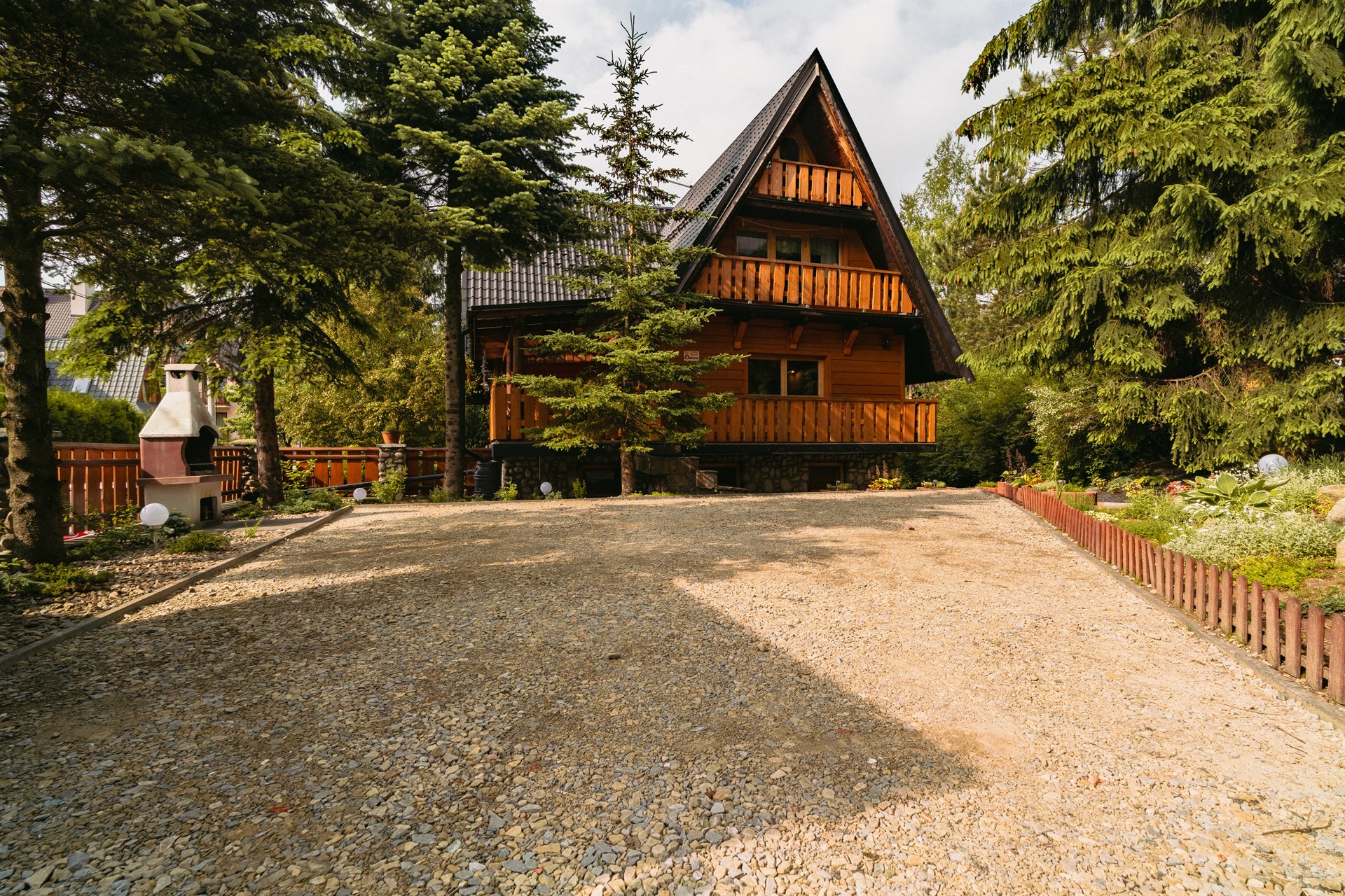 Drewniany domek typu A z balkonem, otoczony choinkami i żwirowym podjazdem, grill po lewej stronie