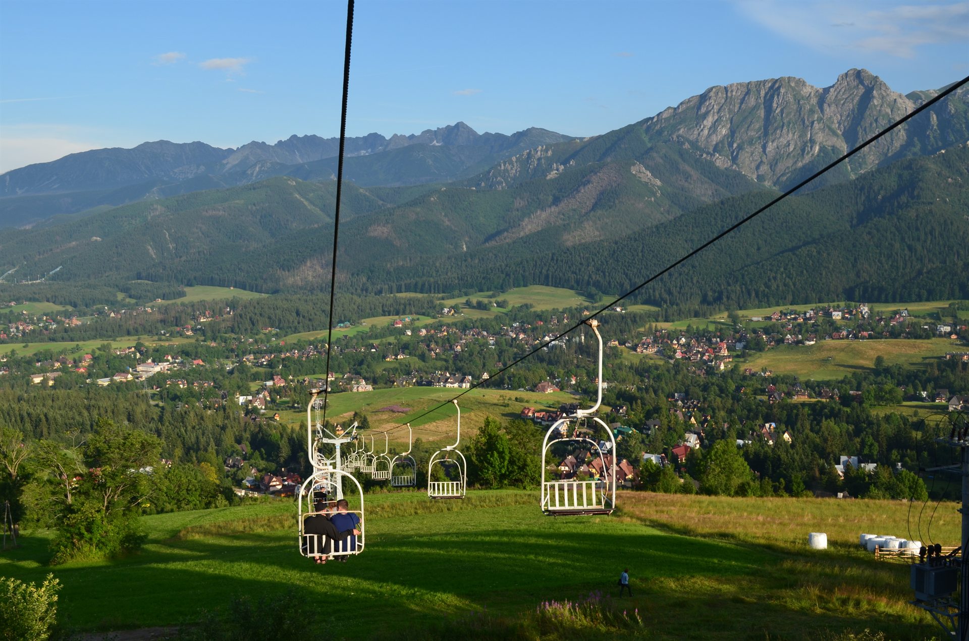 Biały wyciąg krzesełkowy z pasażerami nad zieloną łąką i wsią, w tle wysokie skaliste góry