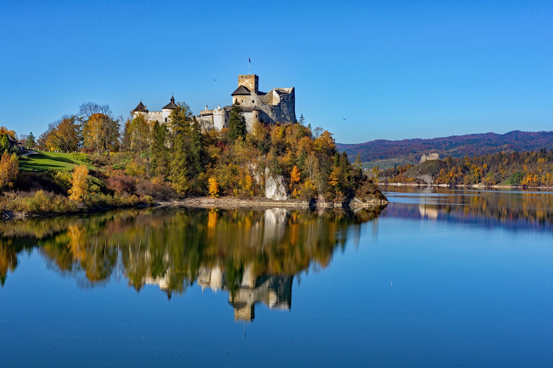 Zamek nad jeziorem na skalistym wzgórzu, jesienne drzewa i idealne odbicie w tafli wody