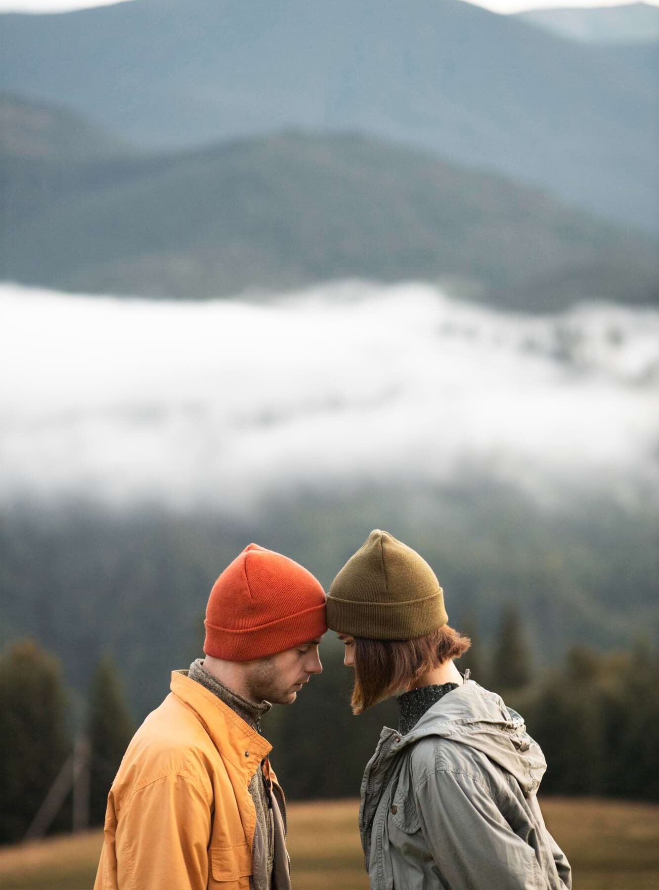 Para w czapkach styka czołami na tle mglistych gór, on w pomarańczowej kurtce, ona w szarej