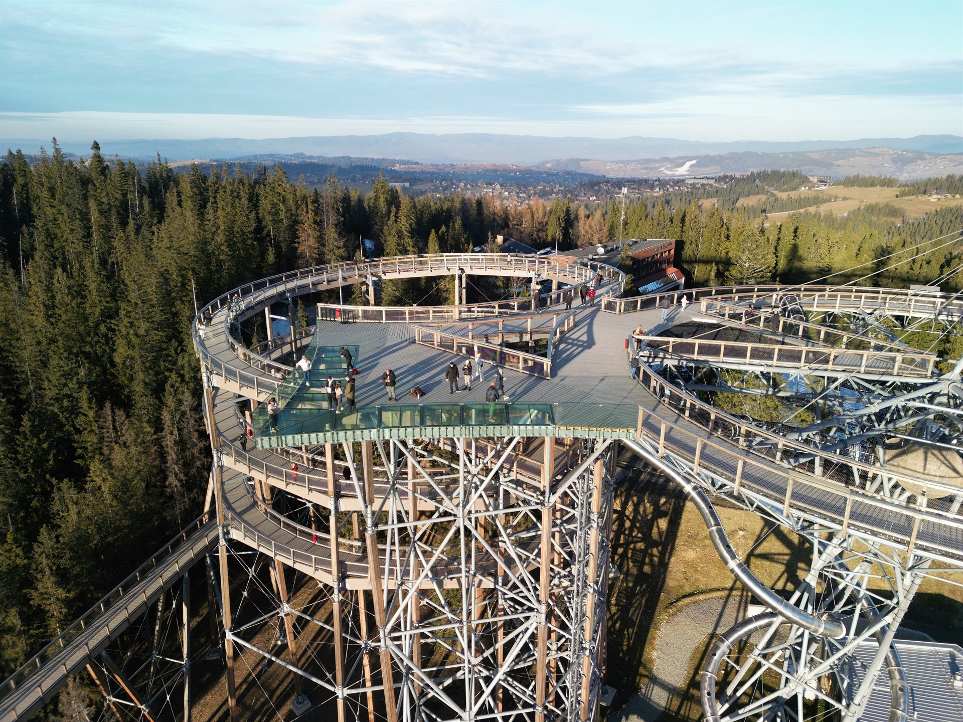 Wysoka drewniano-metalowa platforma spacerowa z szklaną wstawką, spiralnymi chodnikami i rurkową zjeżdżalnią nad lasem