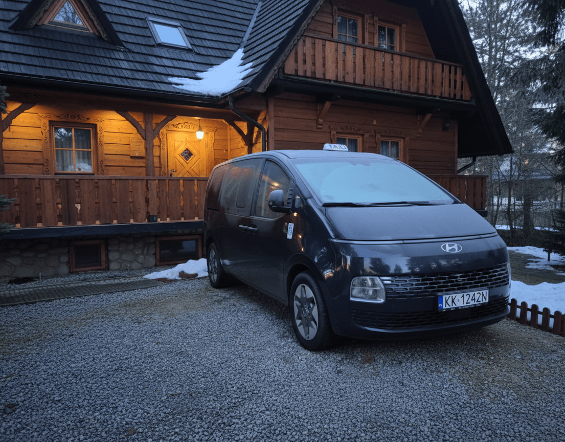 Ciemnoszary van Hyundai z taksówkowym szyldem stoi na żwirowym podjeździe przed drewnianym domkiem z ciepłym światłem