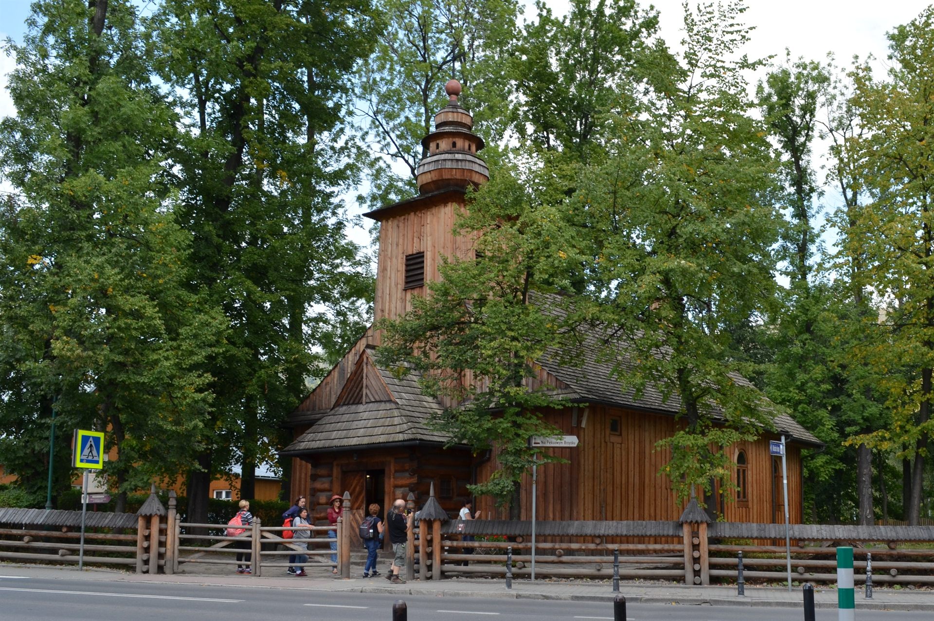 Drewniany kościół z wieżą i gontowym dachem wśród drzew, ogrodzony płotem, z grupą ludzi przy wejściu