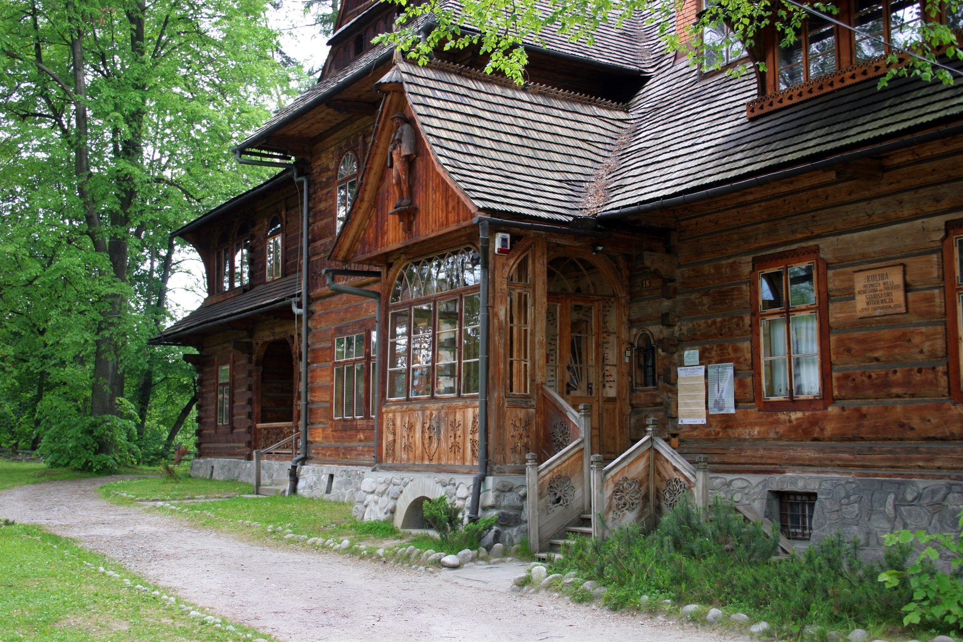 Drewniany dom z rzeźbionym gankiem i gontowym dachem, kamienna podmurówka, przylegająca ścieżka i zielone drzewa